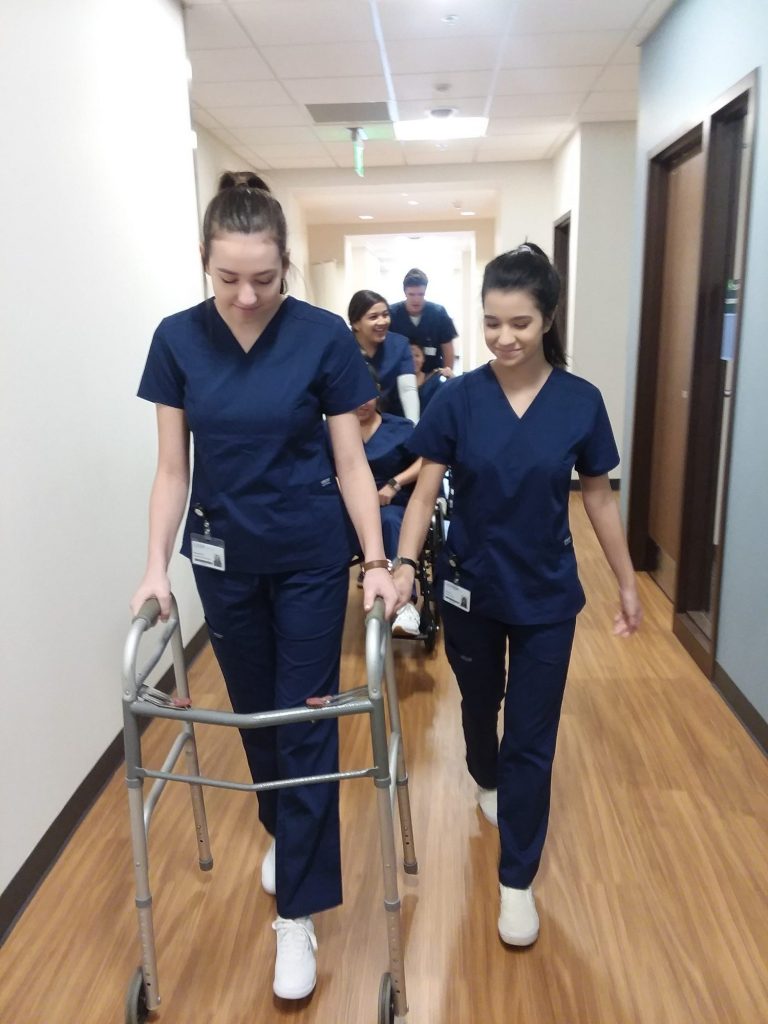 Nurse aide student helping another student walk down a hallway with a walker.